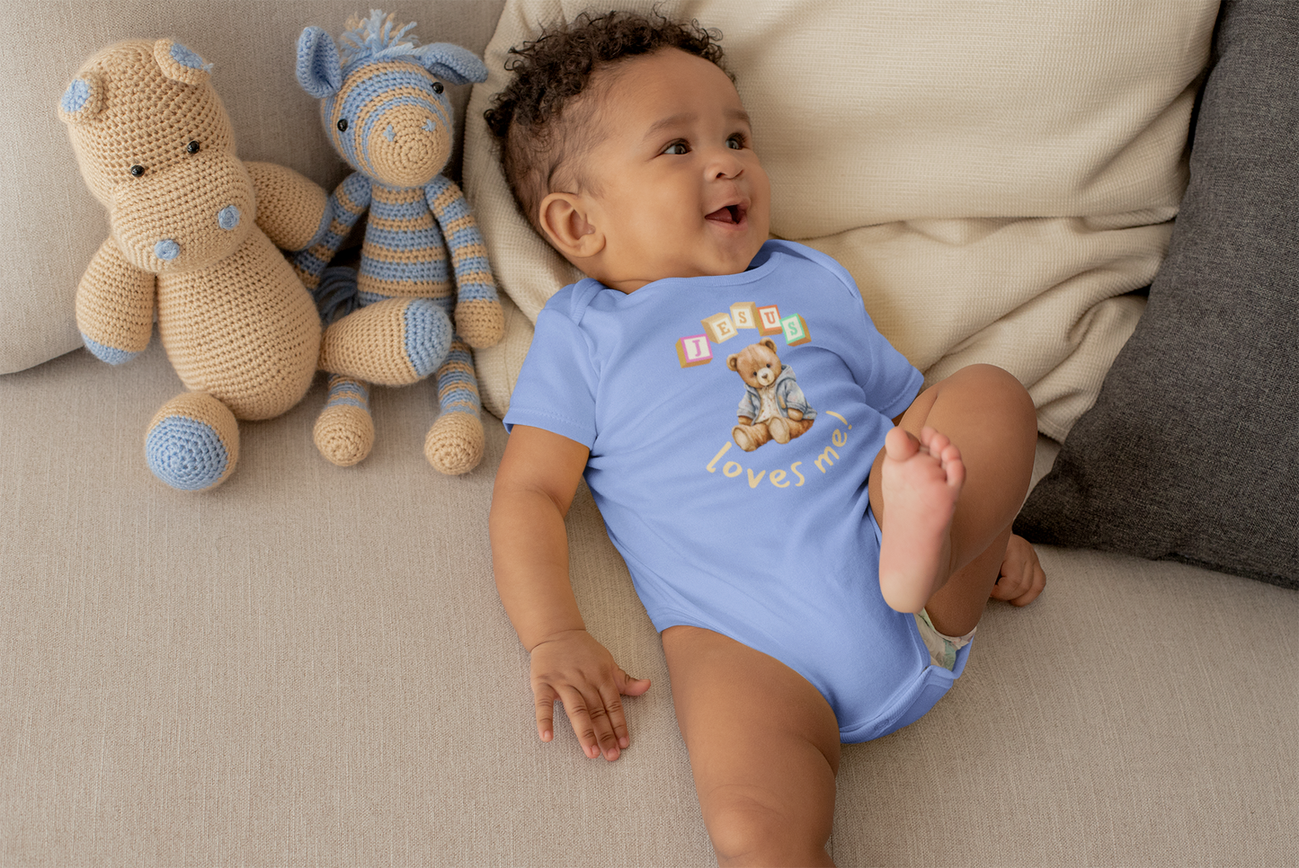 Baby in a blue onesie with teddy bear design sitting on a couch next to stuffed animals.