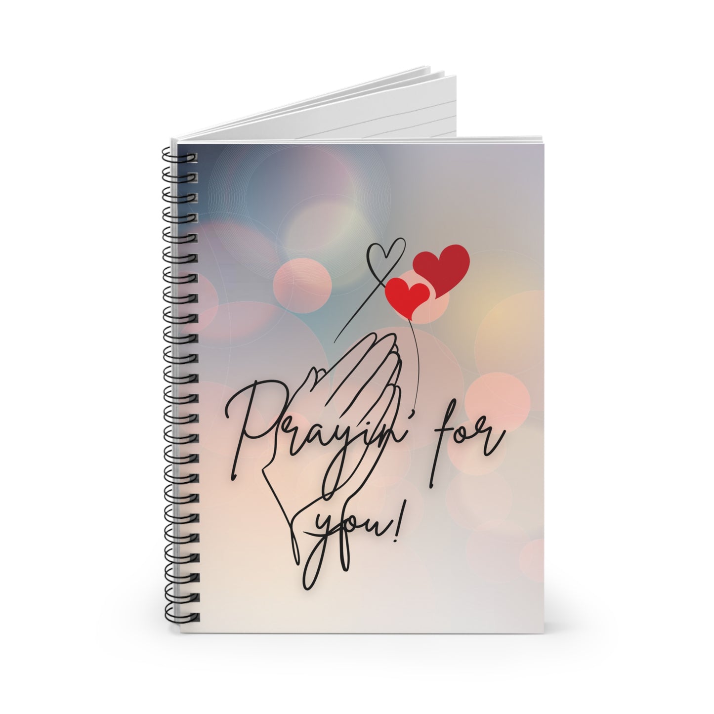 Notebook with a design of hands praying and hearts, text 'Prayin' for you!', on a blurred background.