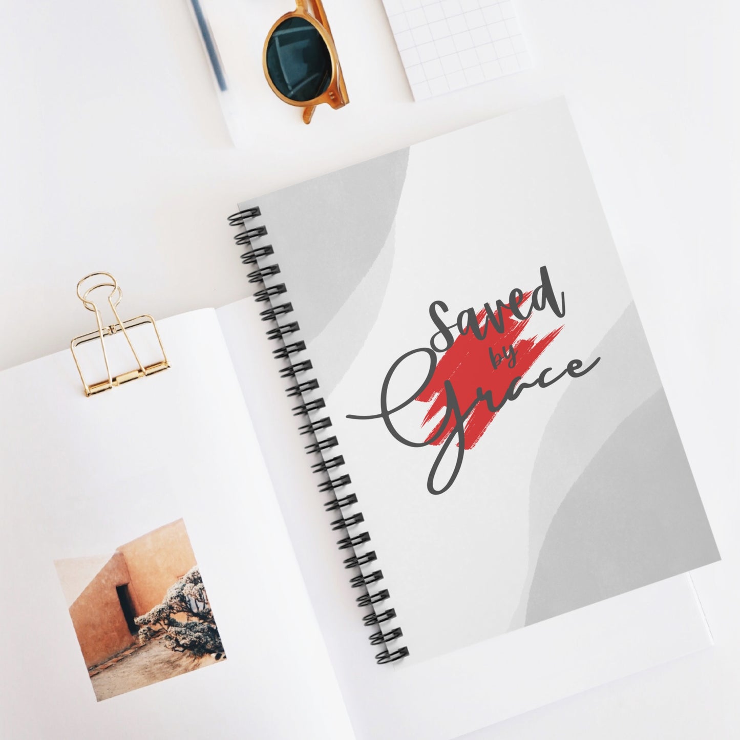 Notebook with 'Saved by Grace' design, photo clip, and sunglasses on a white surface