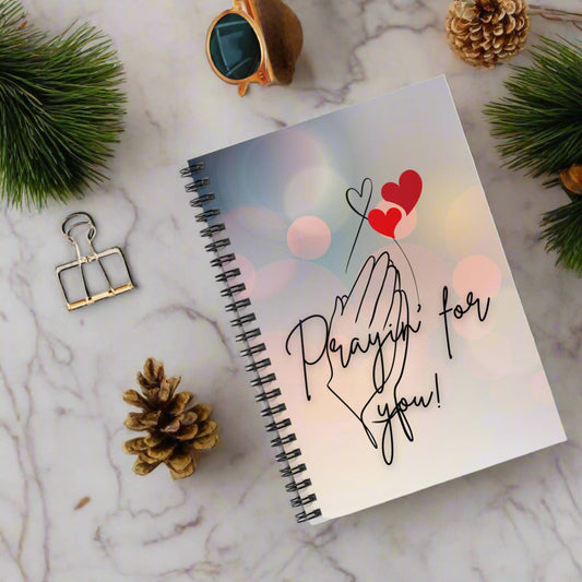 Notebook with 'Praying for you!' text and heart designs on a white surface with sunglasses and a photo clip.