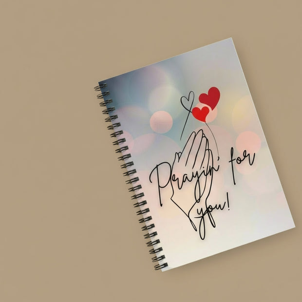 Notebook with 'Praying for you!' text and heart designs on a white surface with sunglasses and a photo clip.