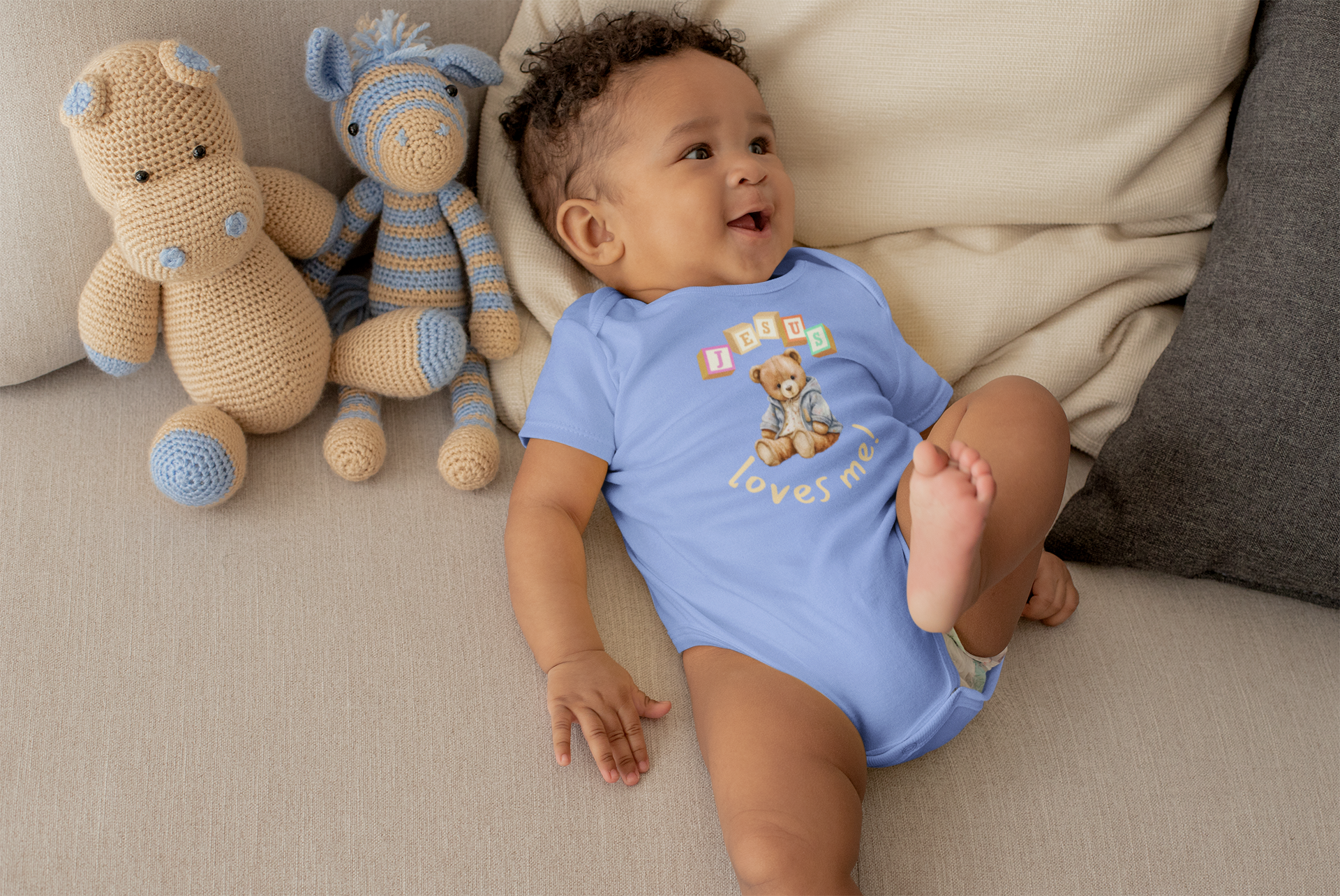 Baby in a blue onesie with teddy bear design sitting on a couch next to stuffed animals.
