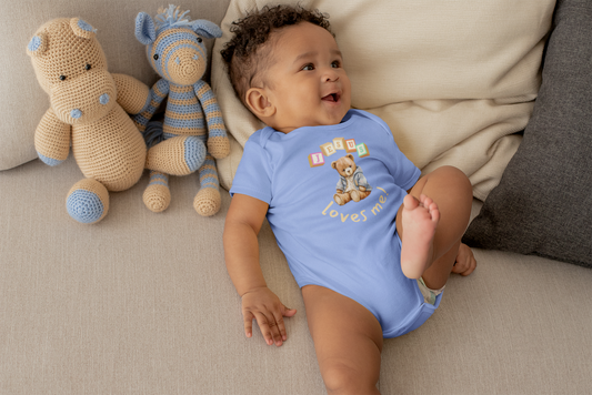 Baby in a blue onesie with teddy bear design sitting on a couch next to stuffed animals.