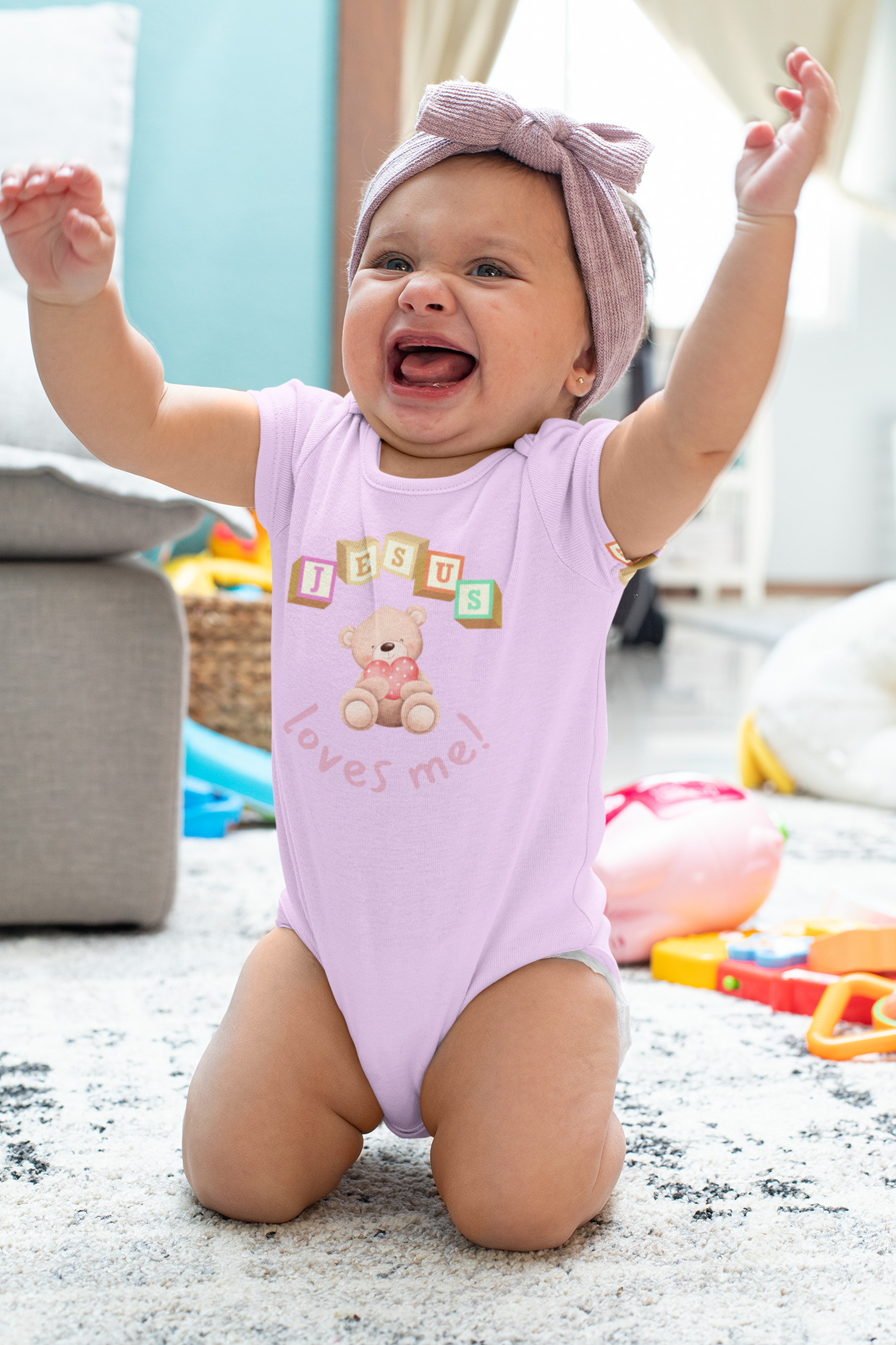 Baby in a pink onesie with teddy bear design and "Jesus Loves Me" text, sitting on a carpeted floor with toys around.