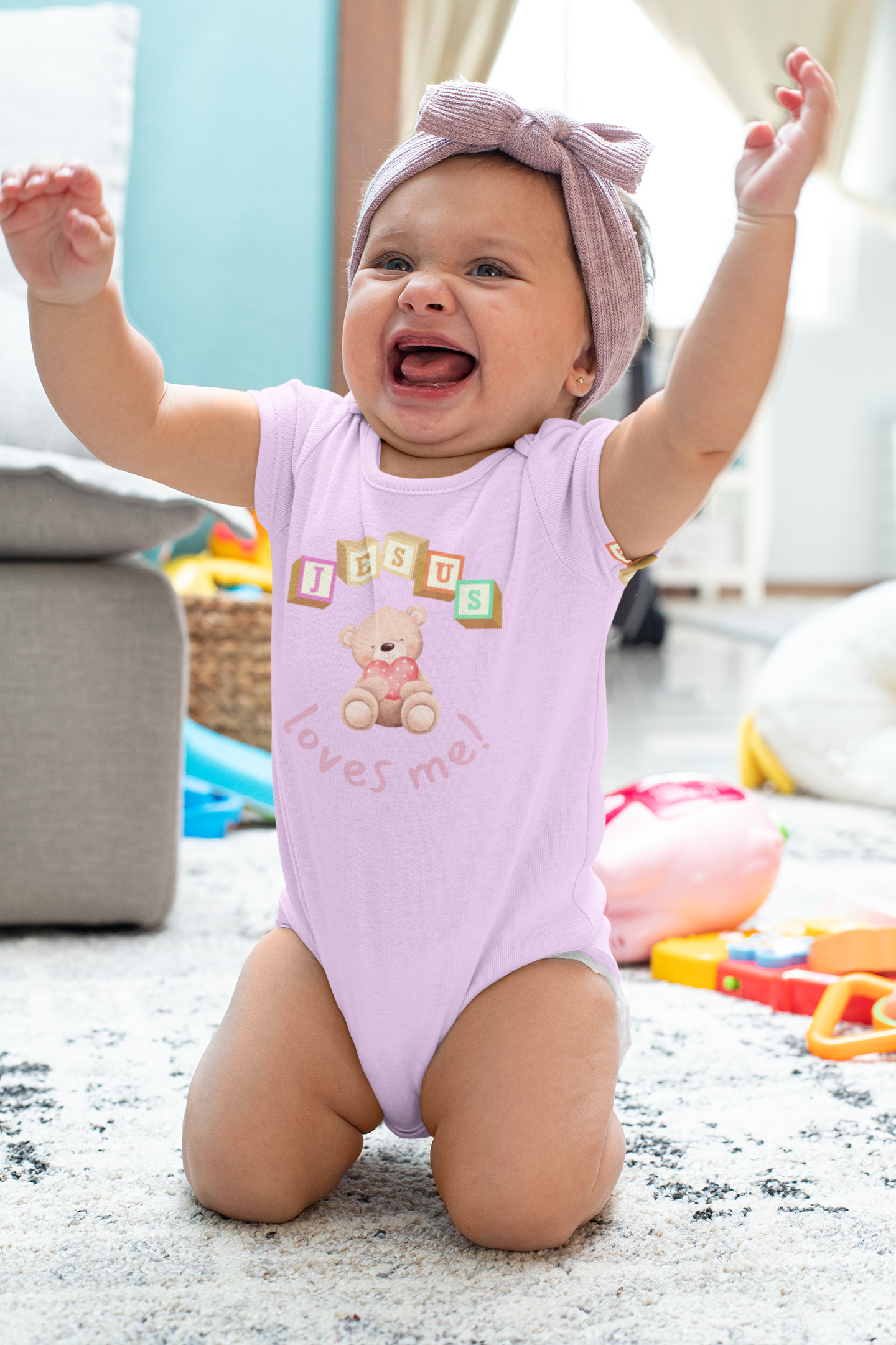 Baby in a pink onesie with teddy bear design and "Jesus Loves Me" text, sitting on a carpeted floor with toys around.