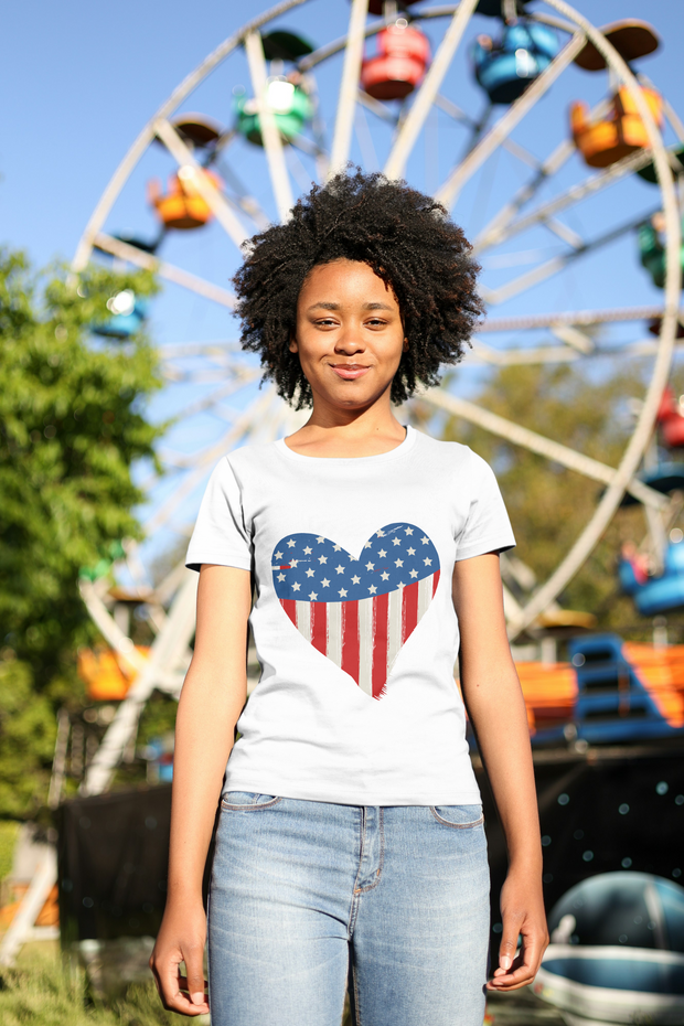 Person wearing a white t-shirt with an American flag heart design in front of a Ferris wheel at an amusement park.